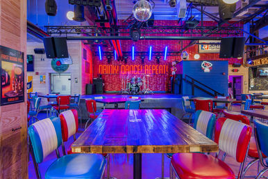 Vibrant Kansas City bar and live-music venue interior with retro red, white and blue diner chairs, wooden tables, disco ball overhead, stage with drum kit and neon “DRINK DANCE REPEAT” sign under blue and purple lighting.