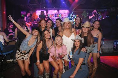 Smiling group of friends in cowboy hats and boots posing with drinks at a lively indoor country bar with a live band on stage, neon lights and balloons.