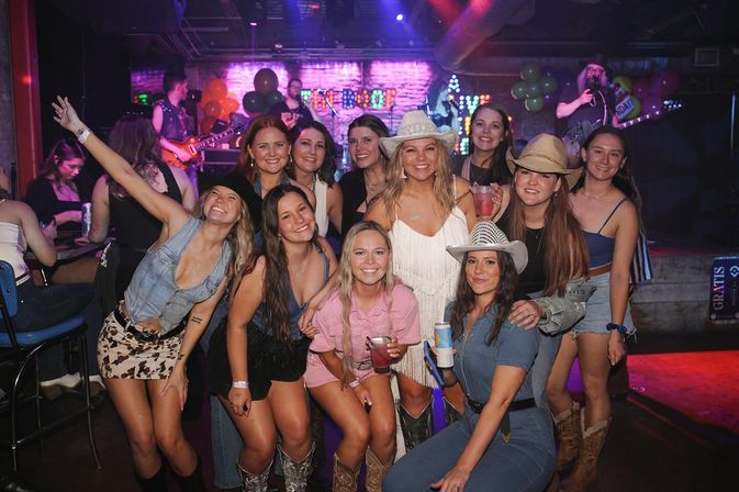 Smiling group of friends in cowboy hats and boots posing with drinks at a lively indoor country bar with a live band on stage, neon lights and balloons.