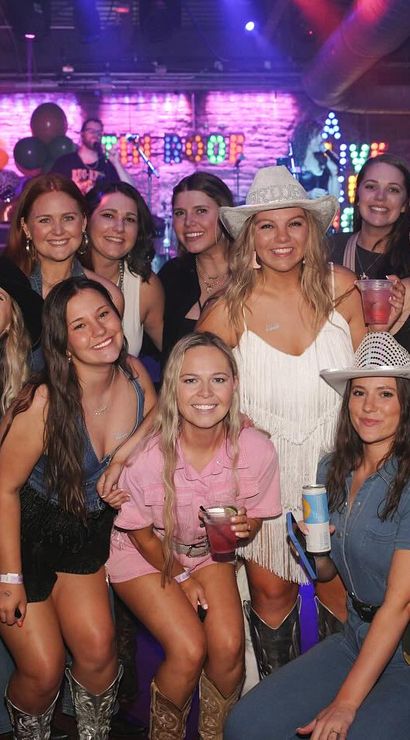 Smiling group of friends in cowboy hats and boots posing with drinks at a lively indoor country bar with a live band on stage, neon lights and balloons.
