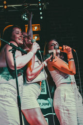 Three women singing into microphones on a dimly lit bar stage during a lively karaoke night