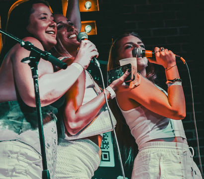 Three friends singing together at a karaoke night on a small indoor stage, holding microphones, a drink and a phone under colorful stage lights with a brick-wall backdrop.