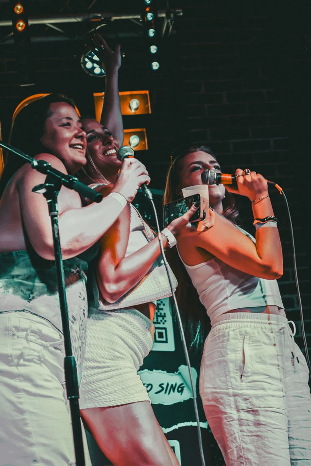 Three friends singing together at a karaoke night on a small indoor stage, holding microphones, a drink and a phone under colorful stage lights with a brick-wall backdrop.