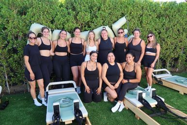 Smiling group of women in black workout gear posing outdoors with Pilates reformer machines on green turf in front of a leafy hedge — sunny group fitness class vibe.