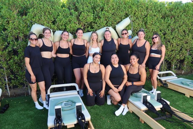 Smiling group of women in black workout gear posing outdoors with Pilates reformer machines on green turf in front of a leafy hedge — sunny group fitness class vibe.