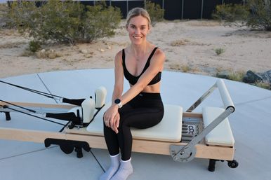 Smiling woman in black sports bra and leggings seated on a Pilates reformer on a concrete pad with desert shrubs — outdoor Pilates workout.