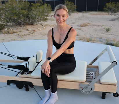 Smiling woman in black sports bra and leggings seated on a Pilates reformer on a concrete pad with desert shrubs — outdoor Pilates workout.