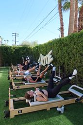 Outdoor reformer Pilates class on green turf under palm trees and a clear blue sky, participants lying on machines with legs raised during a sunny group workout.