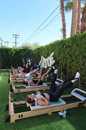 Outdoor reformer Pilates class on green turf under palm trees and a clear blue sky, participants lying on machines with legs raised during a sunny group workout.
