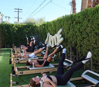 Outdoor reformer Pilates class on green turf under palm trees and a clear blue sky, participants lying on machines with legs raised during a sunny group workout.