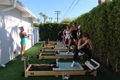 Small-group outdoor reformer Pilates class on wooden machines over artificial turf, women exercising beside a tall hedge with palm trees and a bright blue sky