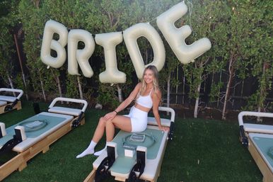 Smiling woman in white activewear sitting on an outdoor Pilates reformer on green turf beneath large metallic 'BRIDE' balloons in a leafy backyard.