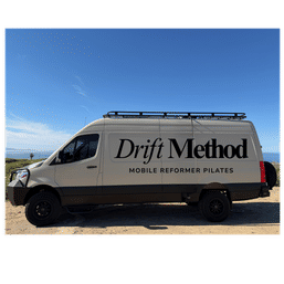 Sunlit coastal scene: beige mobile Pilates van with roof rack and rugged tires parked on a sandy bluff overlooking the ocean, large black lettering advertising mobile reformer Pilates.