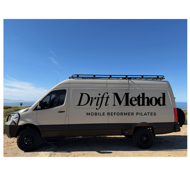 Sunlit coastal scene: beige mobile Pilates van with roof rack and rugged tires parked on a sandy bluff overlooking the ocean, large black lettering advertising mobile reformer Pilates.
