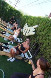 Women in workout gear sipping mimosas during an outdoor Pilates reformer class on artificial grass in a hedged backyard with white inflatable balloons.