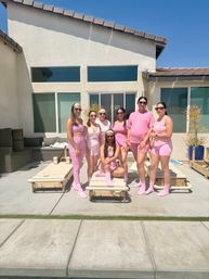 Seven smiling women in matching pink activewear posing on a sunny suburban patio by lounge chairs and a pool in front of a modern stucco house with large windows and blue sky.