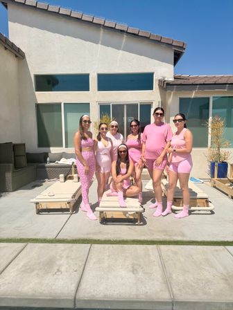 Seven smiling women in matching pink activewear posing on a sunny suburban patio by lounge chairs and a pool in front of a modern stucco house with large windows and blue sky.