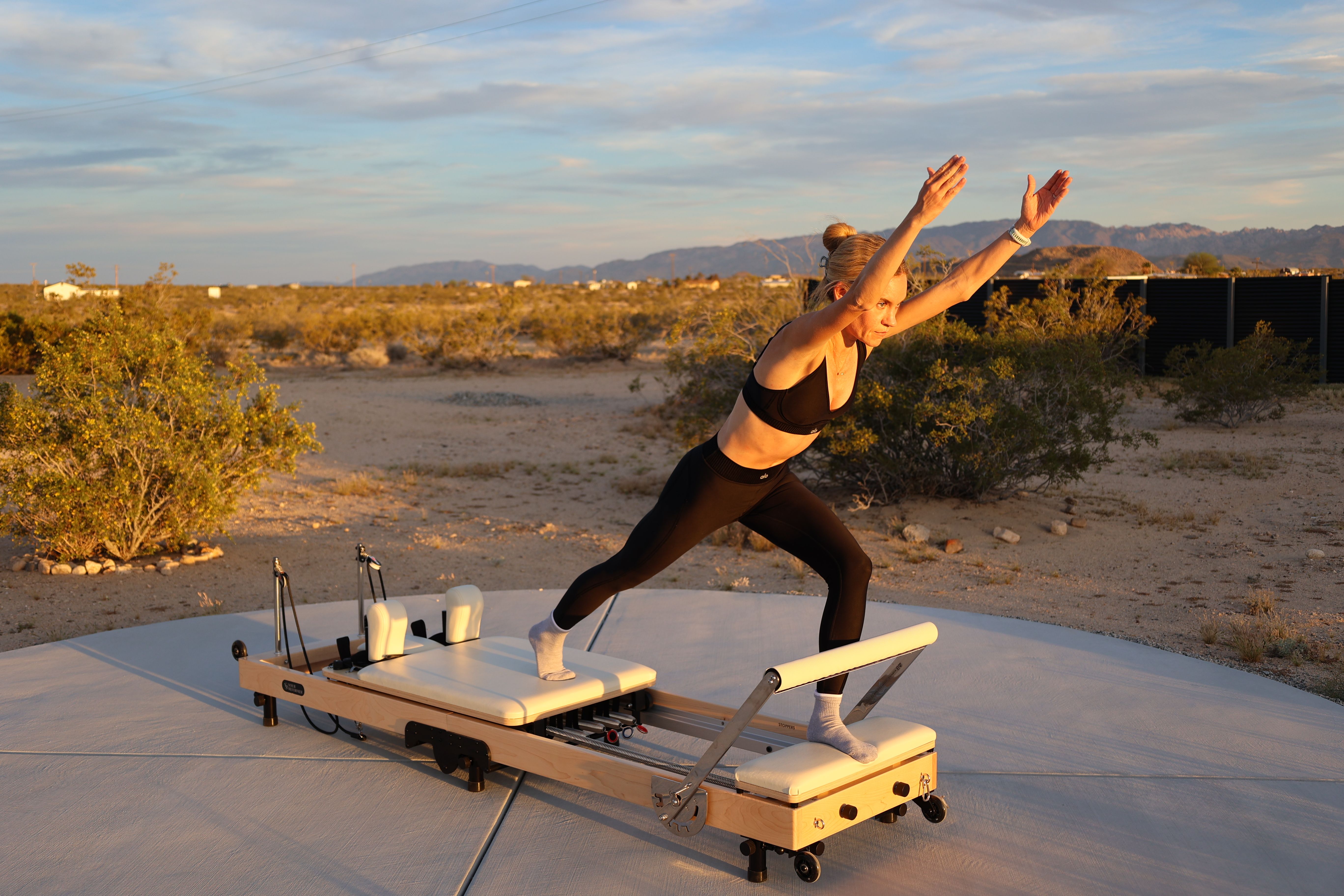 Person performing a Pilates reformer lunge during golden hour on a portable reformer set on a concrete pad in a desert landscape with scrub and distant mountains at sunset.