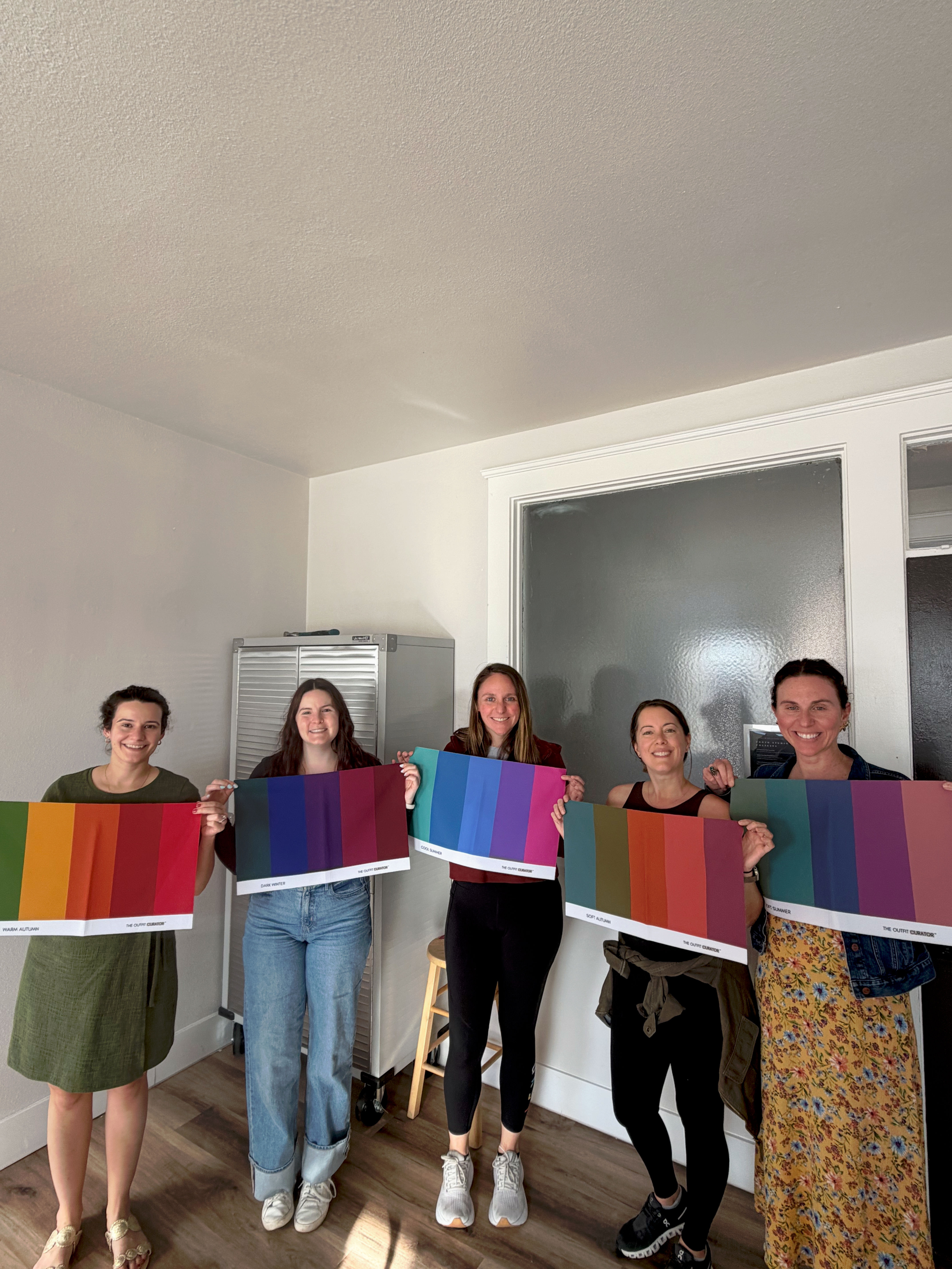 Five women in a bright design studio smiling and holding large vibrant color-swatch boards during an interior design/color consultation workshop, standing on wood floors by a frosted glass door.