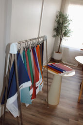 Colorful fabric swatches hanging on a metal sample rack in a sunlit design studio, with stacked samples on a small round table, a potted plant, and wood plank flooring