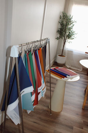 Colorful fabric swatches hanging on a metal sample rack in a sunlit design studio, with stacked samples on a small round table, a potted plant, and wood plank flooring