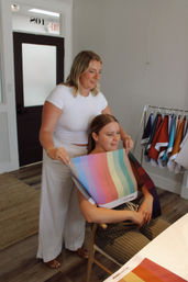 Bright studio color consultation: a stylist drapes a rainbow-striped fabric palette over a seated client's shoulders with a rack of fabric swatches nearby.