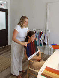 Two women in a bright design studio smiling during a color consultation as one holds a striped fabric swatch against the other, with a rack of color samples and swatch books on a hardwood floor.