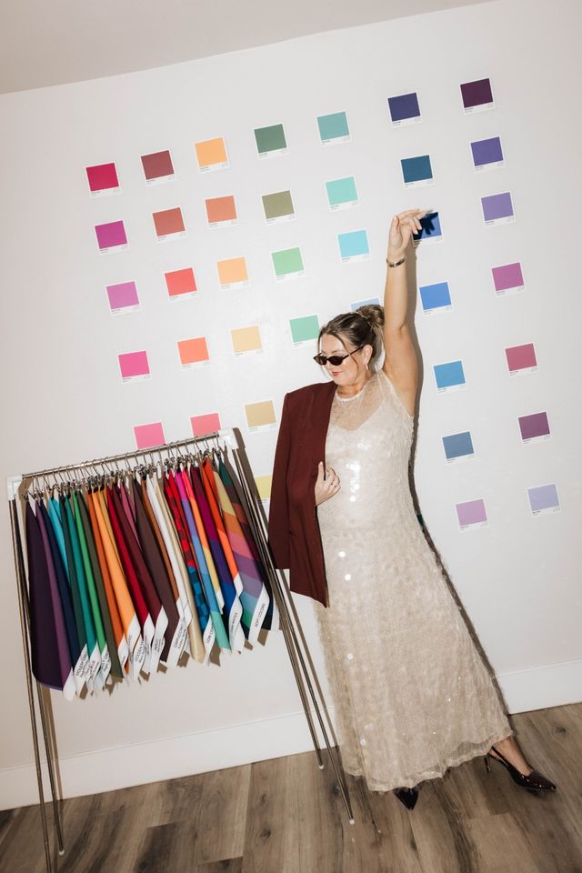 Person in a sequined dress and sunglasses poses in a design studio, draping a maroon blazer next to a rack of vibrant fabric samples and a wall of colorful paint swatches.