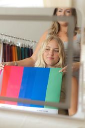 Client smiling in mirror during personal color analysis as a stylist holds bright seasonal swatches (pink, blue, green) in a boutique setting