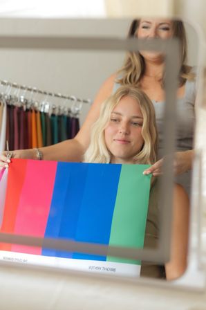 Client smiling in mirror during personal color analysis as a stylist holds bright seasonal swatches (pink, blue, green) in a boutique setting
