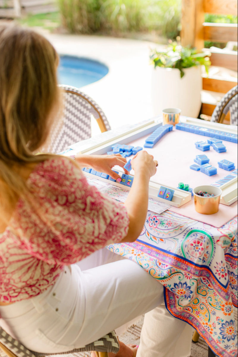 Poolside patio scene: a person arranging bright blue mahjong tiles on a square game table with a colorful patterned tablecloth, small cups with snacks and dice nearby, sunny relaxed vibe.