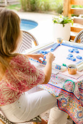 Poolside patio scene: a person arranging bright blue mahjong tiles on a square game table with a colorful patterned tablecloth, small cups with snacks and dice nearby, sunny relaxed vibe.