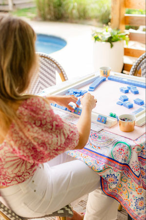 Poolside patio scene: a person arranging bright blue mahjong tiles on a square game table with a colorful patterned tablecloth, small cups with snacks and dice nearby, sunny relaxed vibe.