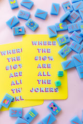 Playful flat-lay of bright yellow cards reading 'WHERE THE #!@% ARE ALL THE JOKERS' surrounded by pastel blue game tiles, small green dice, and a soft pink background