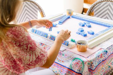 Person's hands arranging blue mahjong tiles on a sunlit automatic mahjong table over a colorful patterned tablecloth, with dice and small cups nearby.