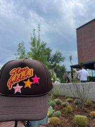 Brown trucker hat with colorful 'Stay Golden!' patch and three stars, held up against an urban patio garden with blurred people, a brick building, and an overcast sky
