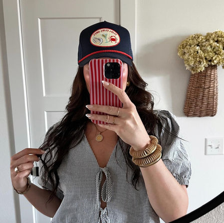 Mirror selfie of a woman in a navy trucker hat with embroidered patch, red-and-white striped phone case hiding her face, gingham tie-front blouse, wavy dark hair, layered necklace and stacked wooden bracelets, wicker wall basket with dried flowers in a bright home