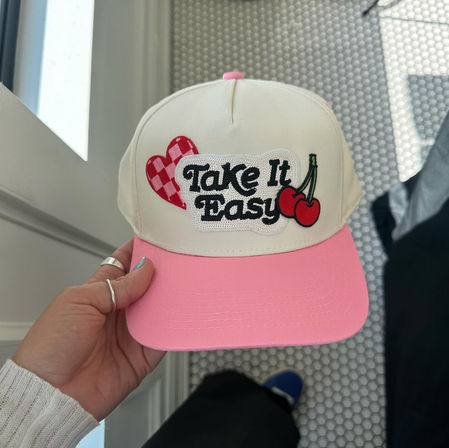 Hand holding a cream-and-pink trucker hat with a pink brim and embroidered "Take It Easy" patch featuring red cherries and a checkered heart, photographed over white hexagon tile flooring.