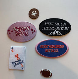 Flat-lay of embroidered iron-on patches on a white background: pink oval "Yappy Hour" patch, black oval "Meet Me on the Mountain" patch with mountain graphic, blue-pink oval "Busy Walking My Dog" patch, small brown paw print circle, playing-card style patch with orange horse and "Best team in Football" text, and a tiny brown football patch.