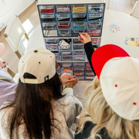 Three people wearing baseball caps sorting colorful stickers and small craft supplies from a clear multi-drawer craft organizer on a white round table