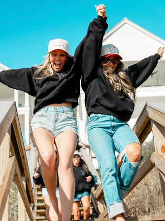 Two friends in black hoodies and caps holding hands and joyfully jumping down wooden beach-house stairs under a bright blue sky, wearing denim shorts and jeans.