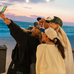 Five friends in caps smiling for a selfie on a sandy beach at sunset, ocean waves and string lights in the background.