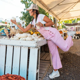 Playful shopper in pink overalls and cap at an outdoor farmers market reaching for a small white pumpkin while holding an iced coffee by a wooden pumpkin display