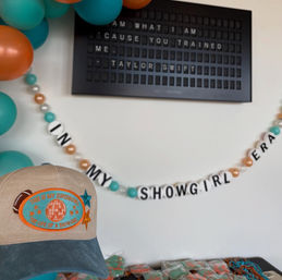 Teal-and-orange party display with balloon cluster, bead-and-letter garland reading 'IN MY SHOWGIRL ERA', a black letter board showing song lyrics, and a tan-and-teal baseball cap with embroidered patch on a table of wrapped treats.