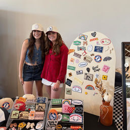 Two smiling women in caps at a pop-up craft market booth featuring a wooden display of colorful embroidered patches and stickers, patch-filled tables, and an orange vase with a white pumpkin accent.