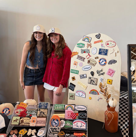 Two smiling women in caps at a pop-up craft market booth featuring a wooden display of colorful embroidered patches and stickers, patch-filled tables, and an orange vase with a white pumpkin accent.