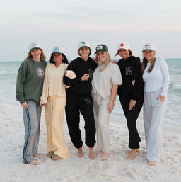 Six friends standing barefoot on a sandy ocean beach at dusk, smiling and wearing casual loungewear and baseball caps in a relaxed seaside group photo