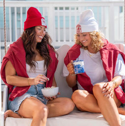 Two smiling friends in beanie hats and red sweatshirts sitting on a sunny porch swing, sharing popcorn from a bowl and a blue patterned mug — casual outdoor deck hangout.