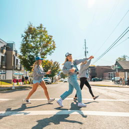 Three friends in casual fall outfits and baseball caps cross a sunny small-town main street at a crosswalk, laughing and holding coffee cups with shops and trees in the background.