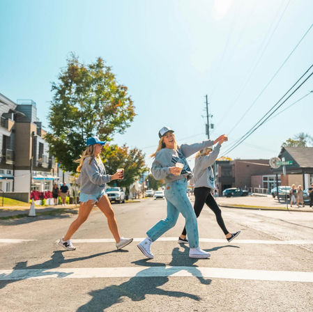 Three friends in casual fall outfits and baseball caps cross a sunny small-town main street at a crosswalk, laughing and holding coffee cups with shops and trees in the background.